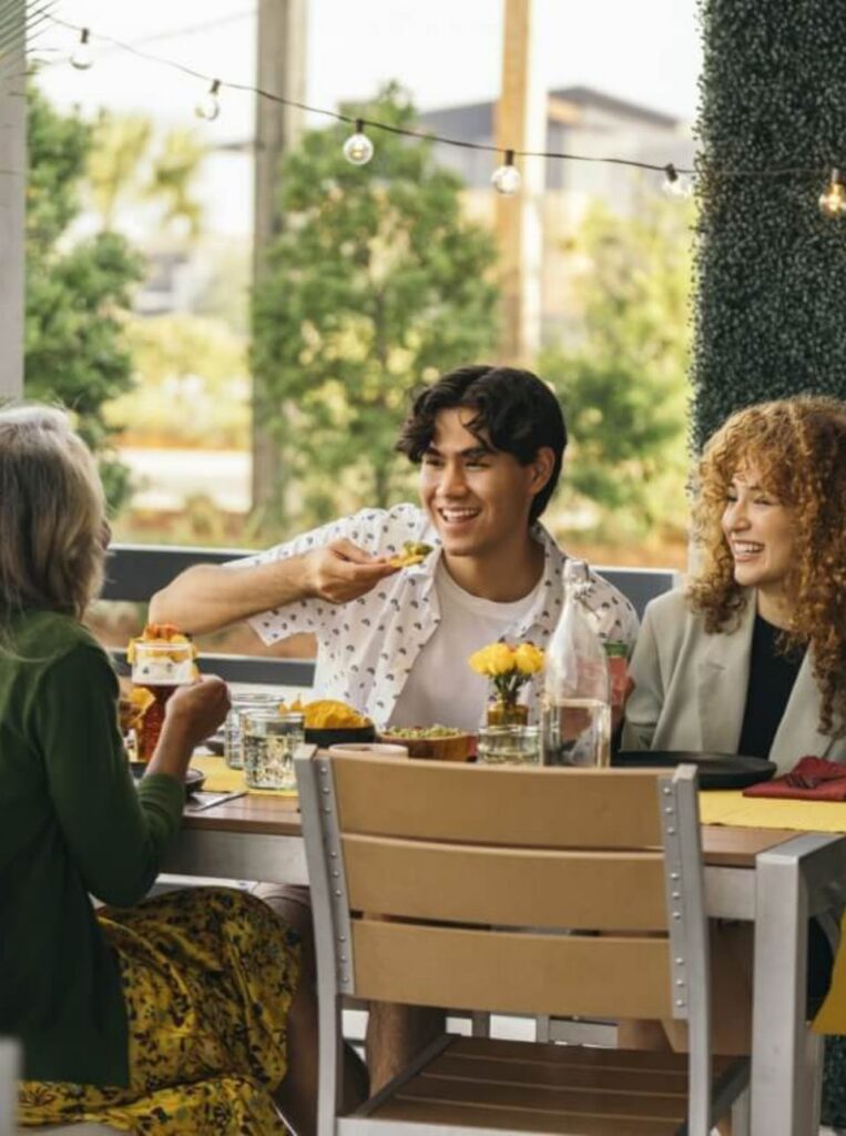 Three people sit around an outdoor table in the Wildlight Yulee Florida community, sharing a meal and lively conversation. Food and drinks are spread out as they enjoy each other's company in this welcoming master planned community.