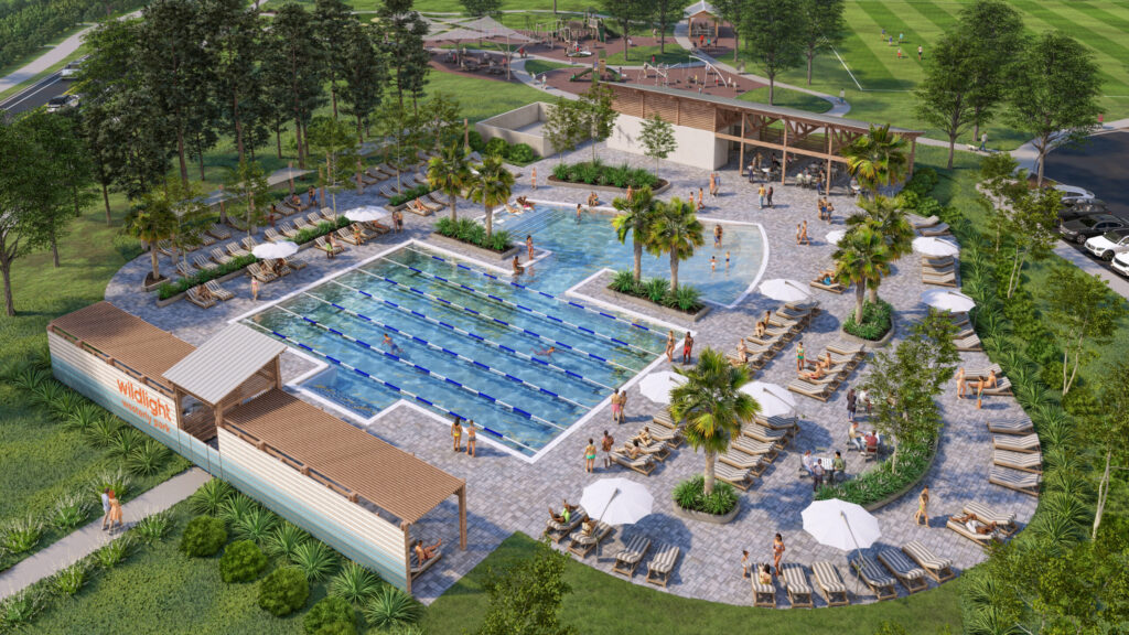 Aerial view of an outdoor community pool area with lap lanes, lounge chairs, umbrellas, palm trees, and people enjoying the Wildlight master planned community; adjacent sports fields and parking lot visible near new homes in Wildlight FL.