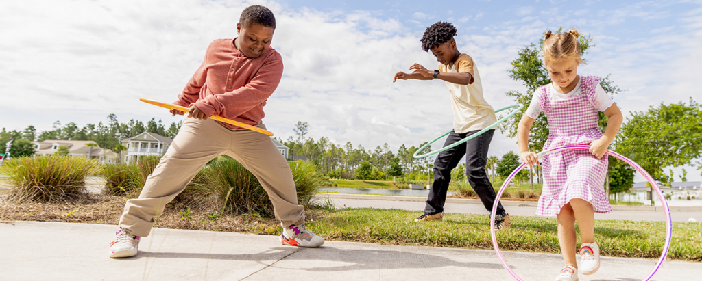 Three children stand outdoors on a sidewalk using hula hoops, with houses and greenery visible in the Wildlight Yulee Florida community under a partly cloudy sky.