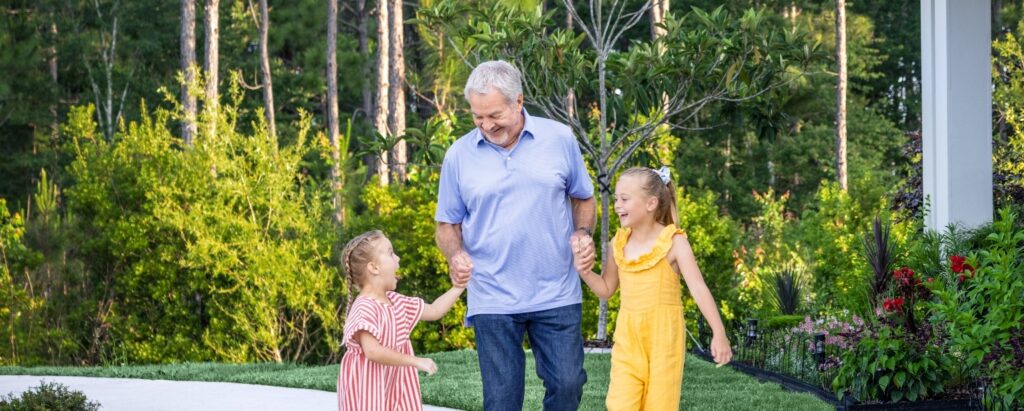 An older man walks hand in hand with two young girls in a garden, all smiling and surrounded by greenery and flowers in the beautiful Wildlight master planned community.