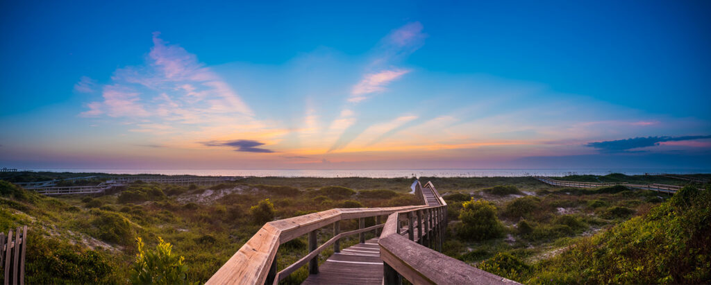 A wooden boardwalk leads through green dunes toward a beach at sunrise in the Wildlight Florida area, with rays of sunlight fanning out across a clear sky.