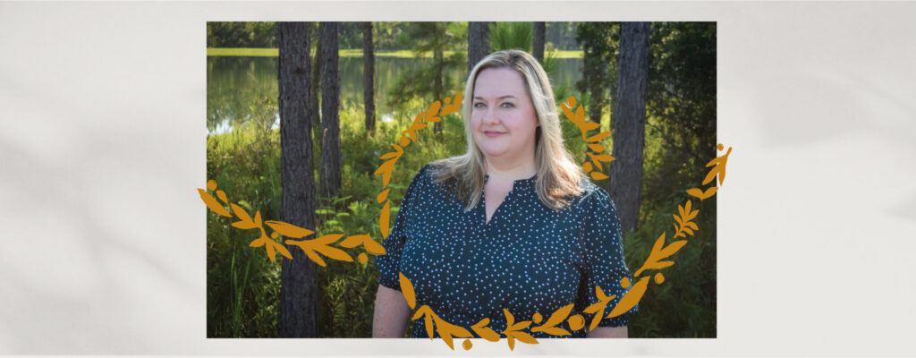 A woman with blonde hair in a dark, patterned shirt stands outdoors near trees and water in the Wildlight Florida community, with an illustrated golden laurel wreath overlay.