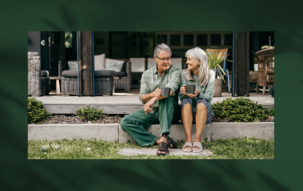 An older man and woman sit together on a concrete ledge outside a house, holding mugs and smiling at each other, surrounded by greenery and patio furniture.