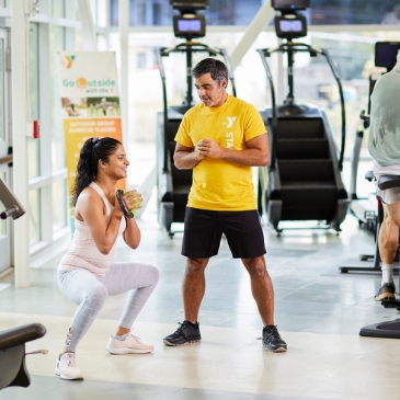 A woman performs a squat with a kettlebell while a male trainer in a yellow shirt observes her in a gym, showcasing the active lifestyle options at Wildlight Florida’s master planned community.