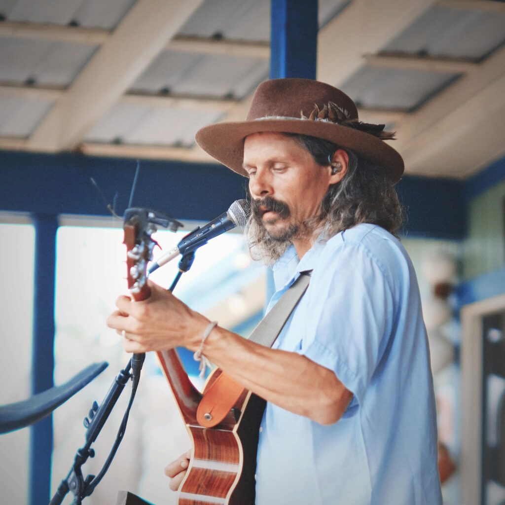 A man with long hair and a mustache, wearing a brown hat and light blue shirt, plays an acoustic guitar and sings into a microphone on an outdoor stage at a Wildlight Yulee Florida community event.