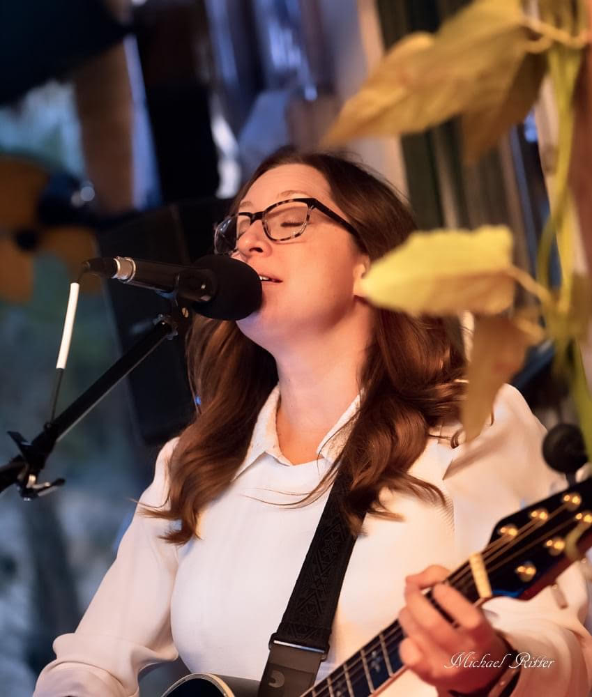 A woman with glasses and long brown hair sings into a microphone while playing an acoustic guitar indoors, sharing her music at a cozy gathering in the heart of the Wildlight Yulee Florida community.