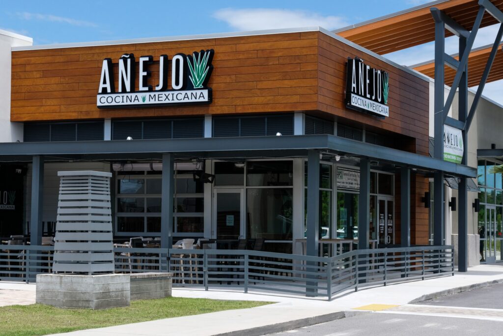 Exterior view of “Añejo Cocina Mexicana,” featuring wood paneling, large windows, and outdoor seating under a partial awning in the vibrant Wildlight Yulee Florida community—perfect for those exploring homes in Nassau County Florida.
