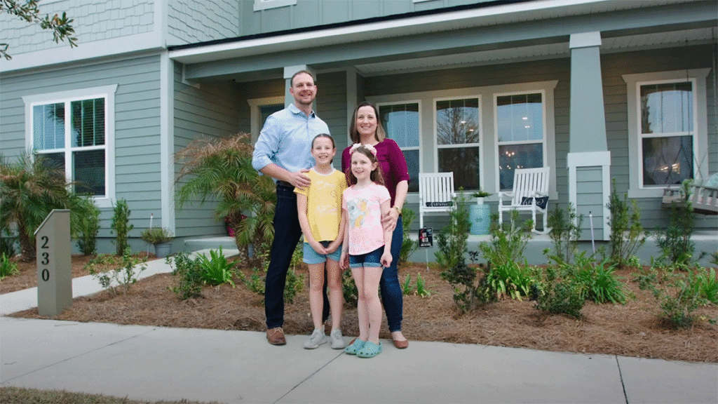 A family of four stands and smiles in front of a two-story house with green siding, white trim, and a small porch, showcasing the warmth of new homes in Wildlight FL.