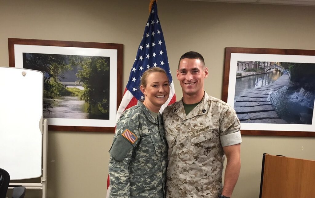 Two people in military uniforms stand smiling in front of an American flag, with framed landscape photos hanging on the wall behind them—reflecting the pride and community spirit found in Wildlight master planned community in Nassau County, Florida.