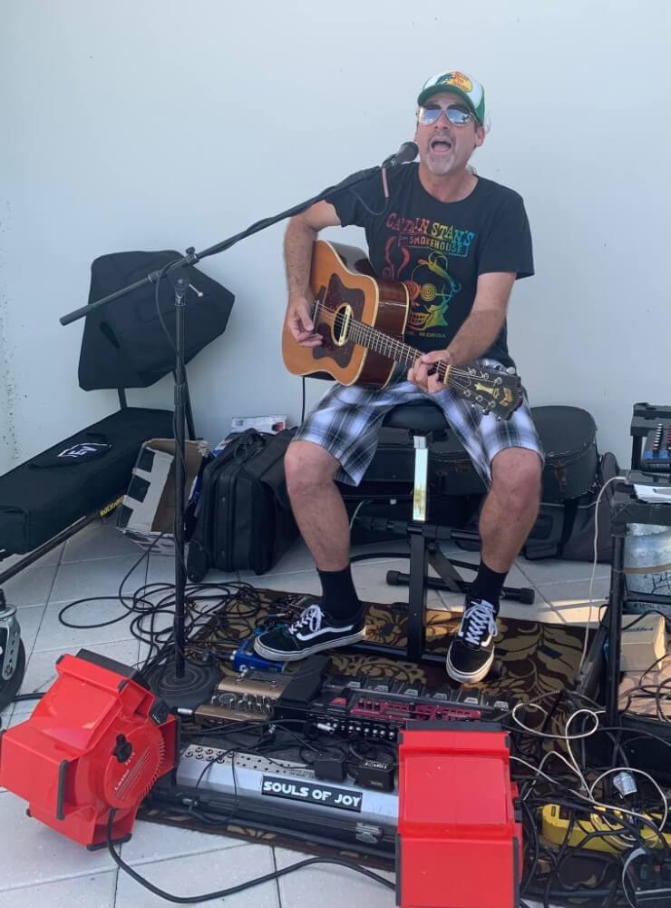 Man sitting and playing an acoustic guitar, singing into a microphone, surrounded by musical equipment and pedals, with a "Souls of Joy" label on the floor—set in the vibrant Wildlight master planned community.