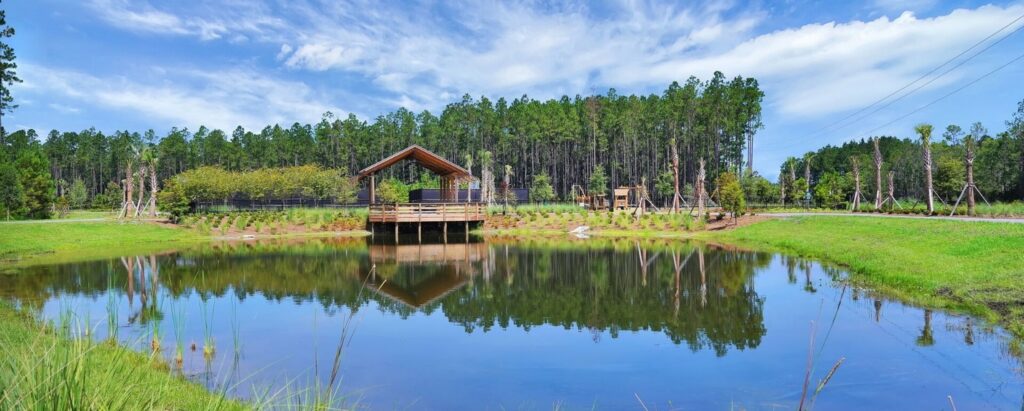 A wooden pavilion sits on the edge of a small pond in the Wildlight Yulee Florida community, surrounded by grass and trees, with its reflection shimmering in the water under a partly cloudy sky.
