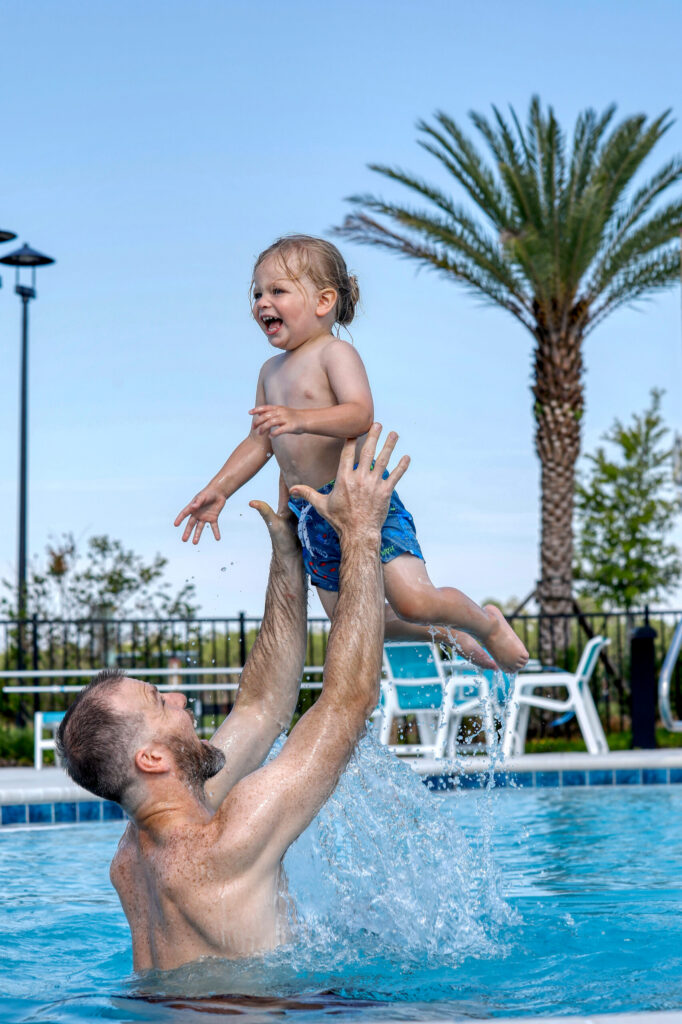 An adult lifts a smiling toddler out of a swimming pool, with water splashing, palm trees, and chairs in the background—capturing the fun family moments possible in the Wildlight master planned community.