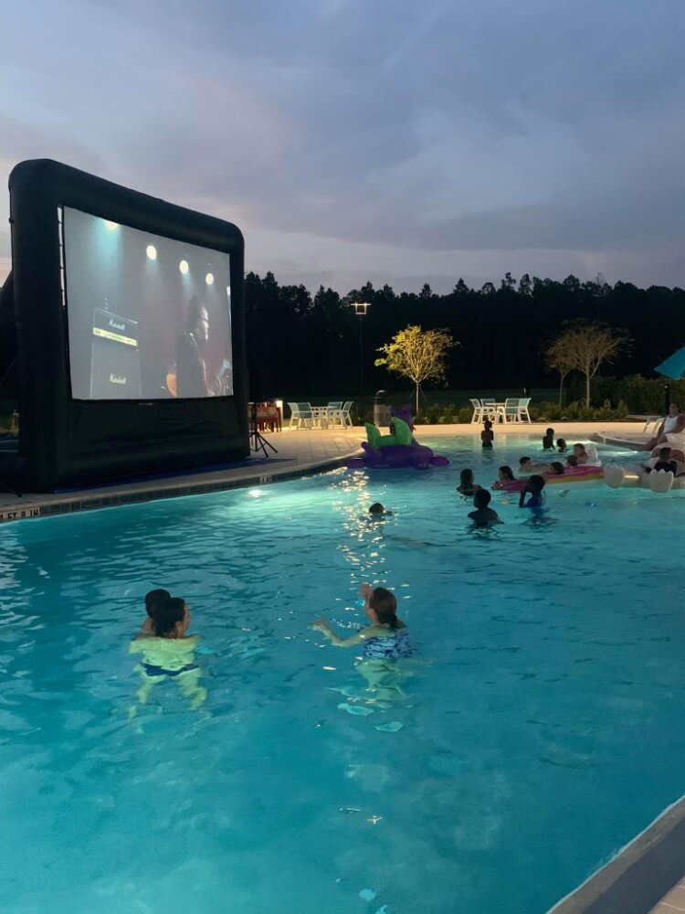 Residents of the Wildlight Yulee Florida community swim in a pool at dusk, enjoying a movie on a large inflatable screen. Some relax on floats while others sit by the water, showcasing the vibrant lifestyle of homes in Nassau County Florida.
