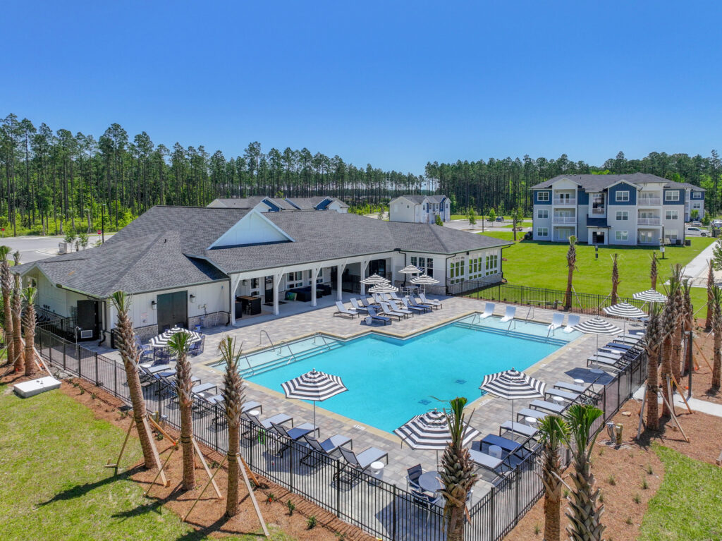 A rectangular outdoor swimming pool with lounge chairs and striped umbrellas, surrounded by a fence; apartment buildings and trees in the background under a clear blue sky in the Wildlight master planned community.