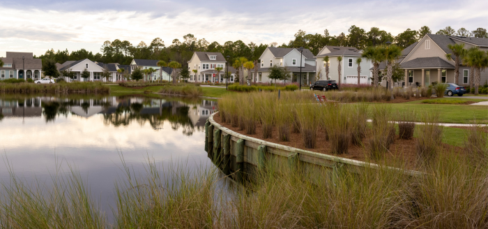Row of suburban houses with garages and parked cars, facing a calm pond with grassy banks and trees in the background under a cloudy sky—typical of new homes in Wildlight FL, part of the vibrant Wildlight Yulee Florida community.