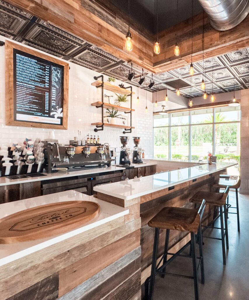 Modern coffee shop interior with wooden accents, bar stools, espresso machines, and a digital menu board—perfect for residents of the Wildlight Yulee Florida community to gather and relax near large windows and enjoy a welcoming atmosphere.
