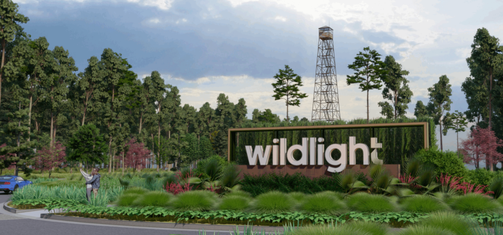 A large "wildlight" sign surrounded by greenery and landscaping sits near a road, welcoming you to the Wildlight master planned community, with a parked blue car, statue, and tall metal structure in the background under a partly cloudy sky.