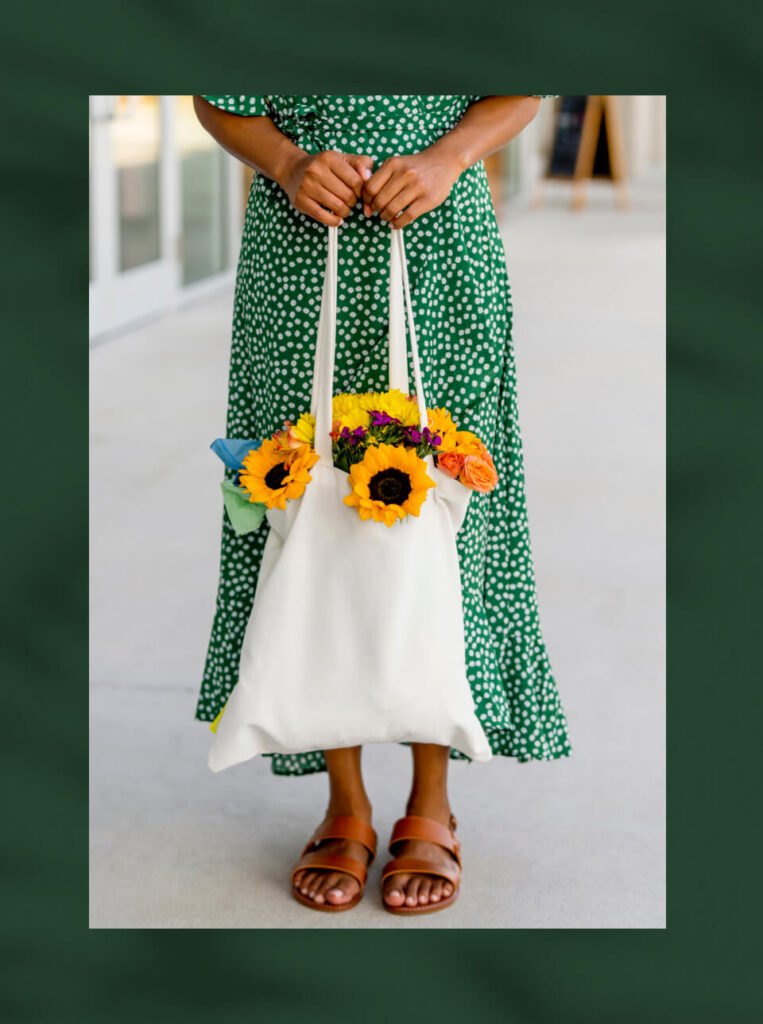 A person in a green polka dot dress holds a white tote bag filled with sunflowers and other colorful flowers, standing on a sidewalk in the vibrant Wildlight Yulee Florida community.