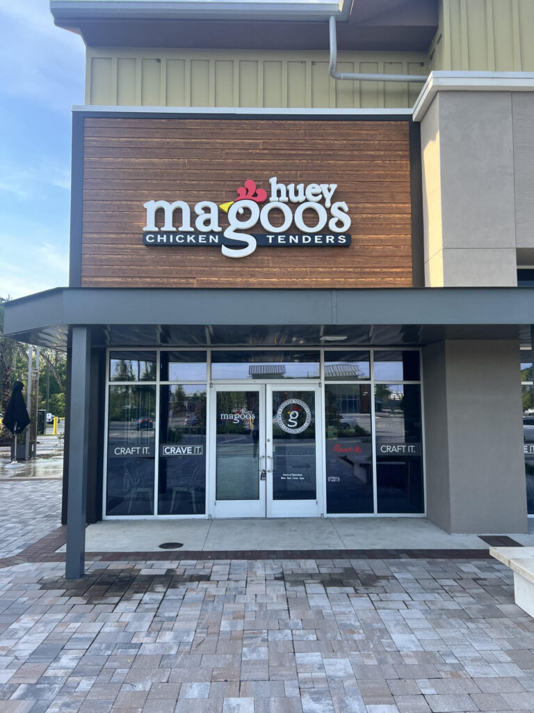 Storefront of Huey Magoo's Chicken Tenders with glass doors and a wooden sign displaying the restaurant's name and logo, welcoming residents and visitors of the Wildlight Yulee Florida community.
