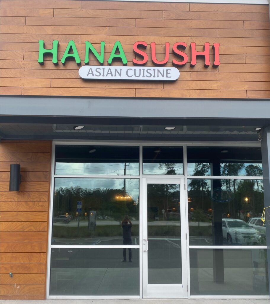 Storefront of Hana Sushi Asian Cuisine with large glass windows and doors, wood panel exterior, and a sign displaying the restaurant's name above the entrance in the heart of the Wildlight Yulee Florida community.