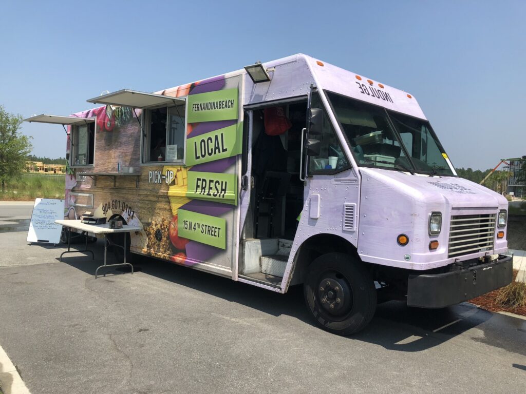 A food truck with a sign reading "Fernandina Beach, Local, Fresh" is parked outdoors near the Wildlight Yulee Florida community, with a table and menu board set up beside it.