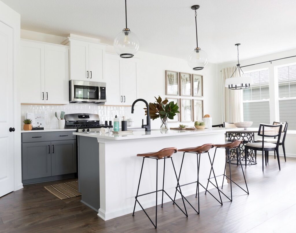 Modern kitchen with a large white island, three brown barstools, stainless steel appliances, pendant lights, and a dining area featuring a round table and large windows—perfect for new homes in Wildlight FL.