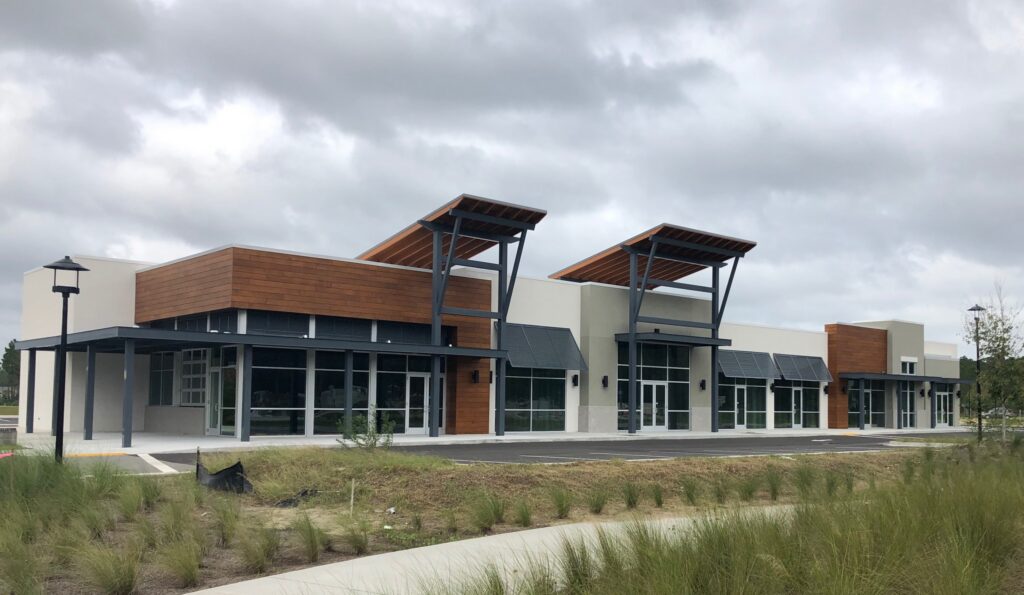 A modern, empty strip mall with large glass windows, wood and metal accents, and an overcast sky in the background sits near homes in Nassau County Florida. The parking lot and sidewalk are visible in front.