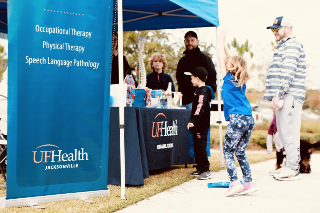 Children and adults interact at a UF Health Jacksonville booth promoting therapy services during an outdoor event in the Wildlight Yulee Florida community. A girl in blue raises her arms, while others watch near the booth.