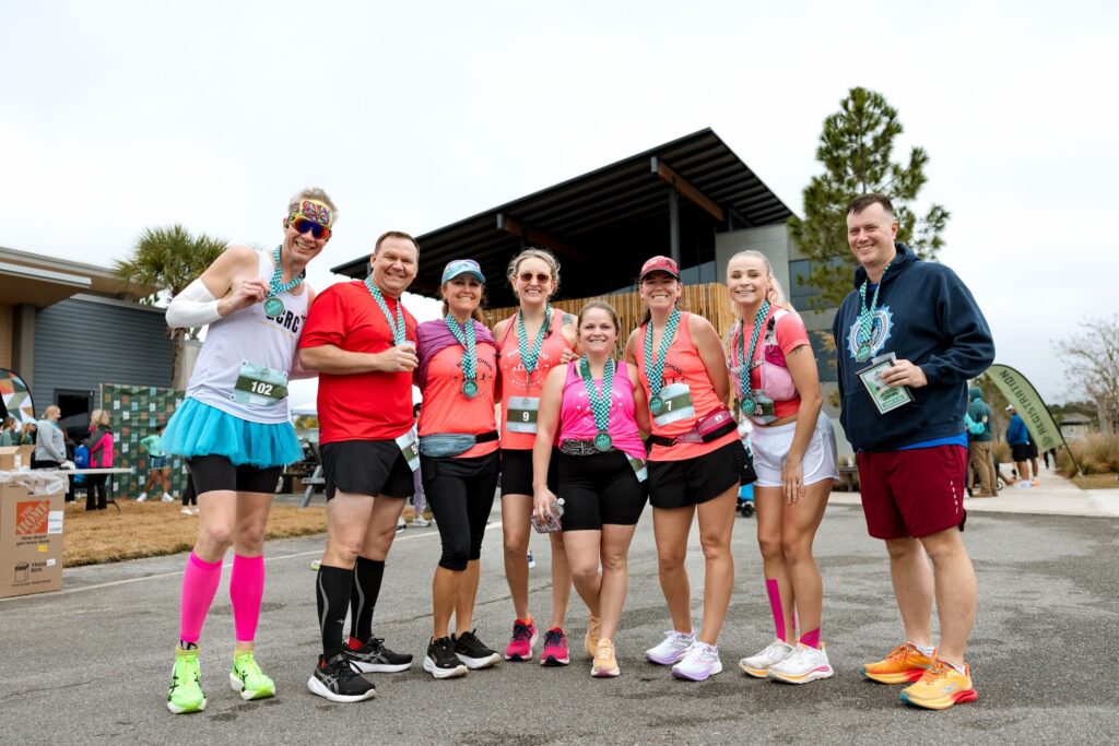 A group of nine runners pose together outside after a race in the Wildlight Yulee Florida community, wearing medals and athletic gear, some in costumes, with a building and trees in the background.