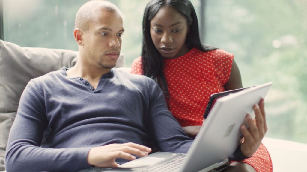 A man and woman sit closely together on a couch in their Wildlight Florida home, looking intently at a laptop screen. The man is typing, while the woman holds a tablet and leans in to see the laptop.