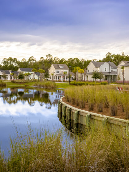 Row of houses beside a small lake with a wooden retaining wall and tall grass in the foreground under a partly cloudy sky, showcasing homes in Nassau County Florida within the inviting Wildlight master planned community.