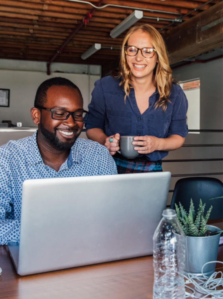 Two coworkers, one seated at a laptop and one standing with a mug, collaborate in a modern office space inspired by the vibrant Wildlight Yulee Florida community, featuring exposed beams and plants.