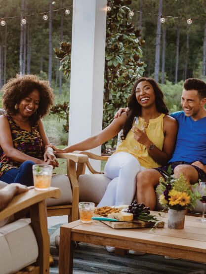 Three people sit on outdoor patio furniture, smiling and holding drinks, with snacks and flowers on a table in front of them. String lights and trees create a cozy atmosphere in the Wildlight Yulee Florida community.