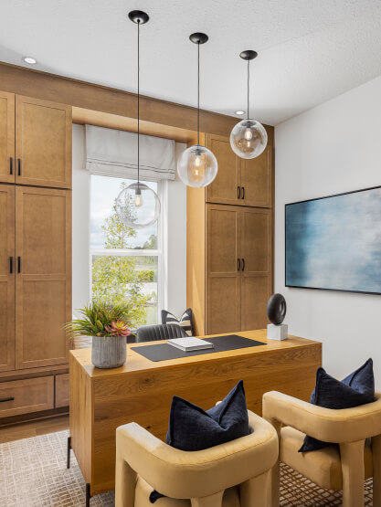 Modern home office with wooden desk, built-in cabinets, two beige chairs, pendant lights, a window with a view, and a large abstract painting—perfect for those seeking new homes in Wildlight FL's vibrant community.