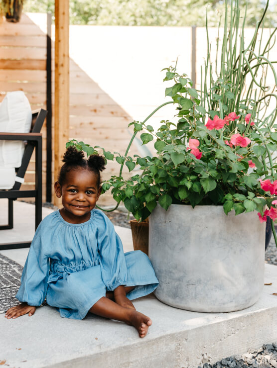 A young child in a blue dress sits barefoot on a concrete step next to a large potted plant with pink flowers, enjoying the outdoors near a wooden fence in the welcoming Wildlight Yulee Florida community.