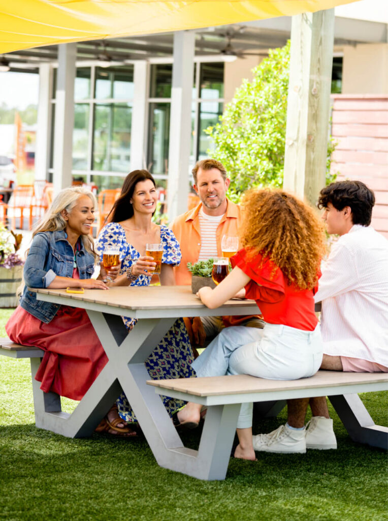 Five people sit around an outdoor table, smiling and conversing with drinks in front of them, enjoying a sunny day in the Wildlight Yulee Florida community.