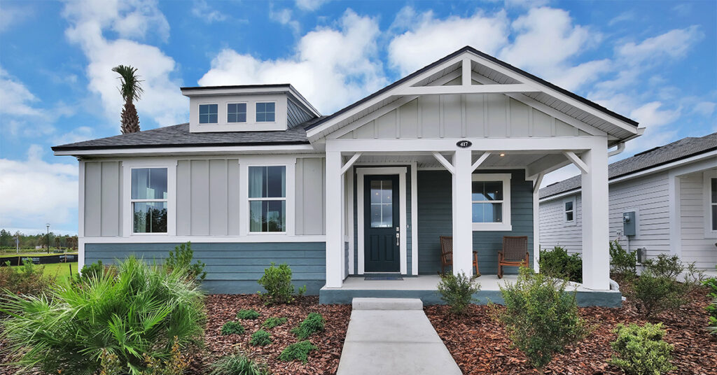 Single-story suburban house with light gray siding, a covered front porch, two chairs, mulch landscaping, and a concrete walkway—an inviting example of new homes in Wildlight FL under a blue sky with scattered clouds.