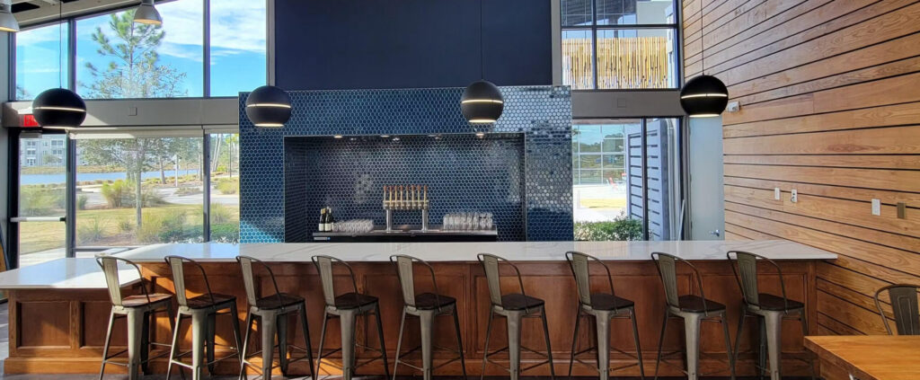 A modern kitchen with a long wooden island, metal barstools, pendant lights, and a tiled backsplash beside large windows—perfect for entertaining in your new home in the Wildlight Yulee Florida community.