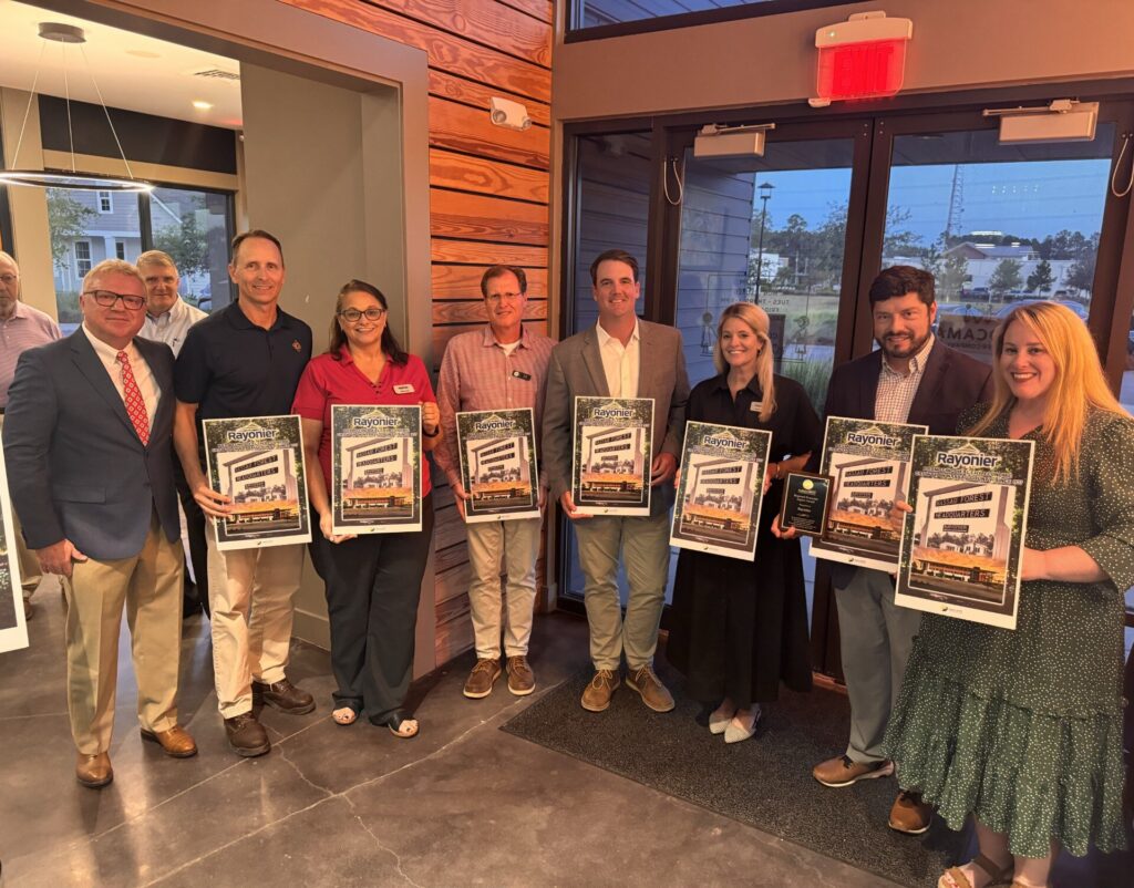 A group of eight people stand indoors, each holding a framed poster with a building design from the Wildlight Yulee Florida community, while one person also holds a plaque. They appear to be at an award or recognition event.