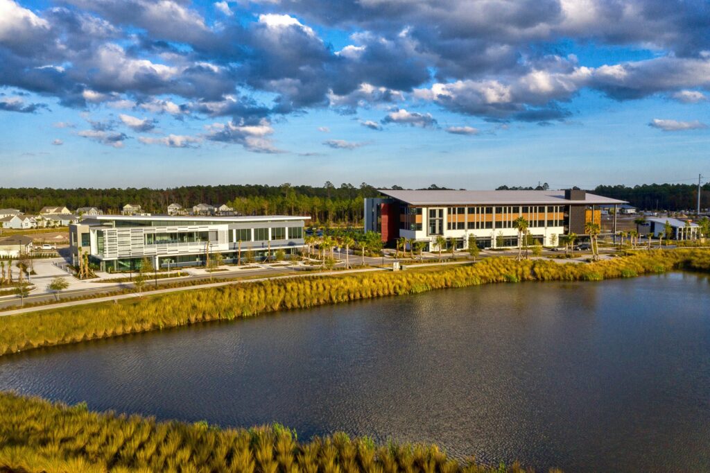 Two modern office buildings sit beside a pond, surrounded by landscaped greenery in the Wildlight master planned community, with homes in Nassau County, Florida and a forest visible under a partly cloudy sky.