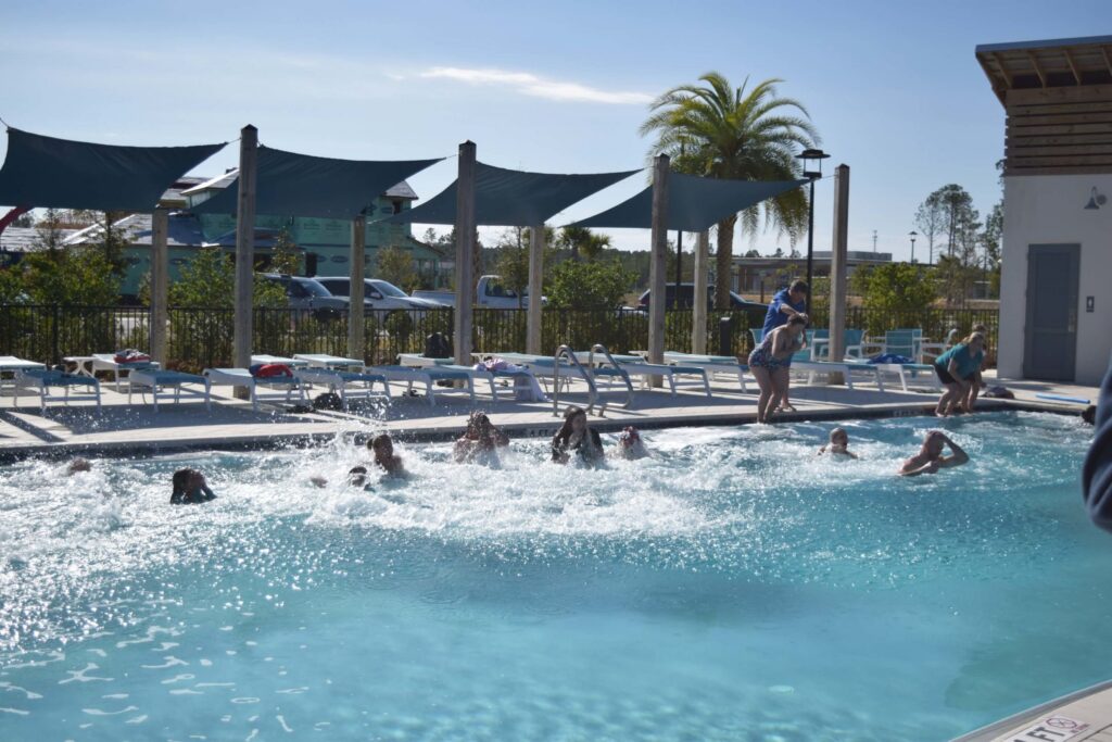 People swim and splash in an outdoor pool under sun shades at Wildlight master planned community, with lounge chairs and palm trees in the background on a sunny day.
