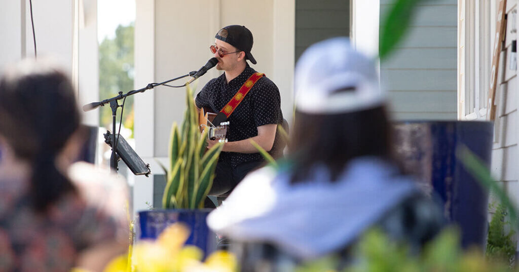 A musician wearing sunglasses and a backwards cap plays guitar and sings into a microphone outdoors at a Wildlight Yulee Florida community event, with an audience seated in the foreground.