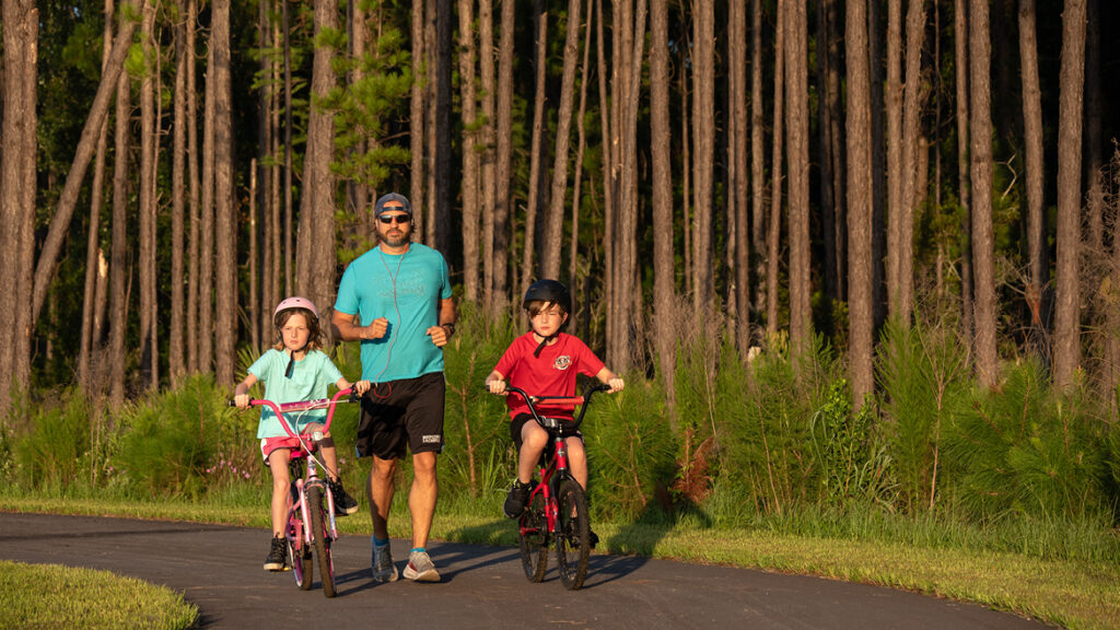 An adult jogs on a paved path beside two children riding bicycles, surrounded by tall trees and greenery in the Wildlight Yulee Florida community—an area known for scenic trails and beautiful new homes in Wildlight FL.