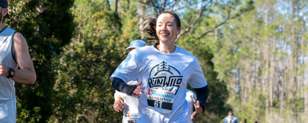 Woman wearing a race bib and "Run Wild" t-shirt runs outdoors on a sunny day, smiling, with other runners in the background—capturing the active spirit of life at Wildlight master planned community.