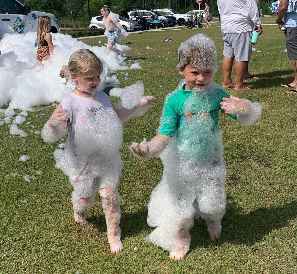Two children covered in foam bubbles walk on the grass at a lively Wildlight Florida outdoor event. Other people and playful foam fill the background, capturing the fun-filled spirit of the Wildlight master planned community.