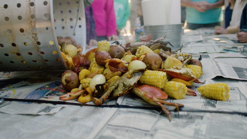 A pile of boiled crabs, corn, potatoes, and sausage is dumped onto a newspaper-covered table at an outdoor gathering in the welcoming Wildlight Yulee Florida community, with people enjoying the feast in the background.