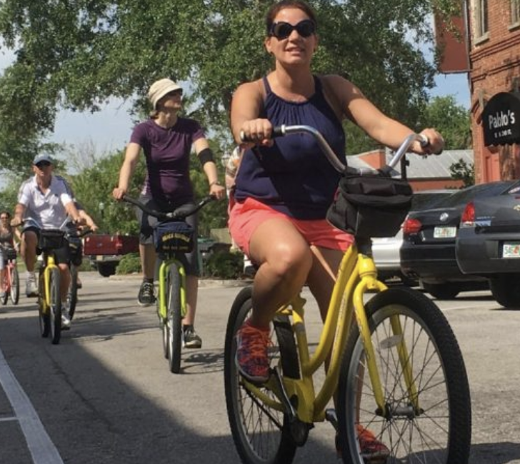 Three people ride bicycles on a sunny street in the Wildlight Yulee Florida community, with trees, parked cars, and a brick building in the background.