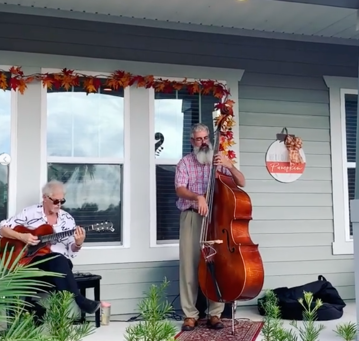 Two men, one playing an acoustic guitar seated and the other on upright bass standing, perform on a porch with autumn leaves and a "Hello Pumpkin" sign in the heart of Wildlight Yulee Florida community.
