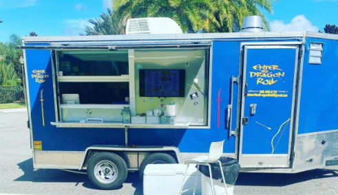 A blue food truck labeled "Enter Dragon Roll" is parked outdoors in the Wildlight Yulee Florida community, its service window open with a menu and various items inside. A white chair and small table sit in front, inviting residents of new homes in Wildlight FL to enjoy.