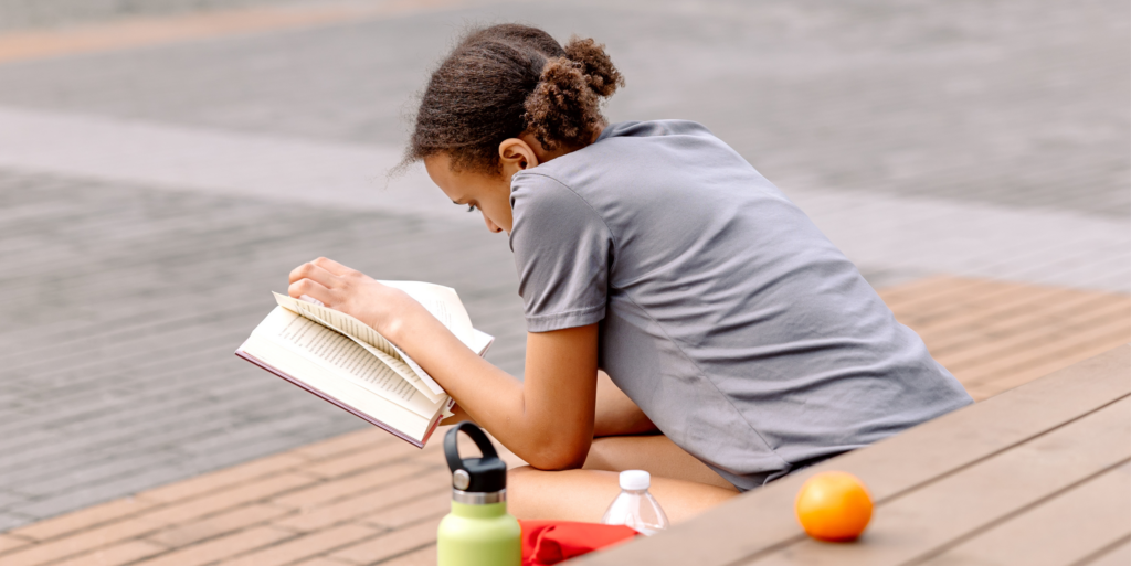 A person sits on a bench outdoors in the Wildlight Yulee Florida community, reading a book, with a water bottle, an orange, and a green bottle beside them.